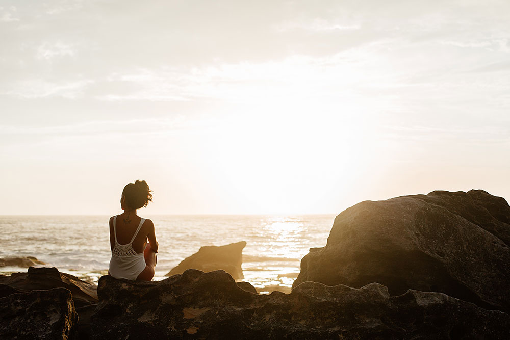 Eine Frau sitzt nachdenklich bei Sonnenuntergang am Meer und blickt in die Ferne